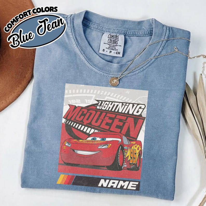 May include: A blue short-sleeved t-shirt featuring a graphic print of the red race car Lightning McQueen. The design includes the text "LIGHTNING MCQUEEN" and "NAME" above a tri-color stripe. The shirt has a Comfort Colors brand label.