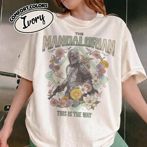 May include: An ivory t-shirt featuring a graphic of The Mandalorian and Grogu surrounded by a floral wreath. The text reads THE MANDALORIAN and THIS IS THE WAY. A small oval label in the top left corner displays the text COMFORT COLORS IVORY.