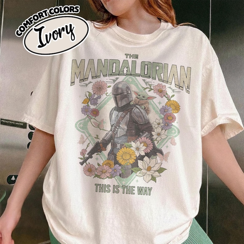 May include: An ivory t-shirt featuring a graphic of The Mandalorian and Grogu surrounded by a floral wreath. The text reads THE MANDALORIAN and THIS IS THE WAY. A small oval label in the top left corner displays the text COMFORT COLORS IVORY.