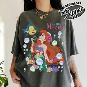K&ouml;nnte beinhalten: Ein anthrazitfarbenes Kurzarm-T-Shirt mit einer Grafik von Arielle und Fabius aus Arielle, die Meerjungfrau. Das Design zeigt bunte Unterwasserelemente wie Blasen, Korallen und Muscheln sowie den Schriftzug Arielle, die Meerjungfrau in Pink.