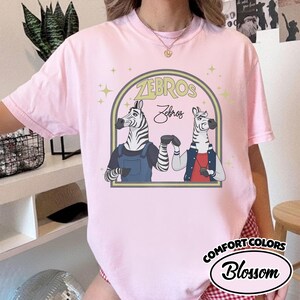May include: A pale pink t-shirt with a cartoon illustration of two zebras inside an arch. The zebras are dressed in outfits, with the word "ZEBROS" above them. The shirt also has the text "Comfort Colors Blossom".