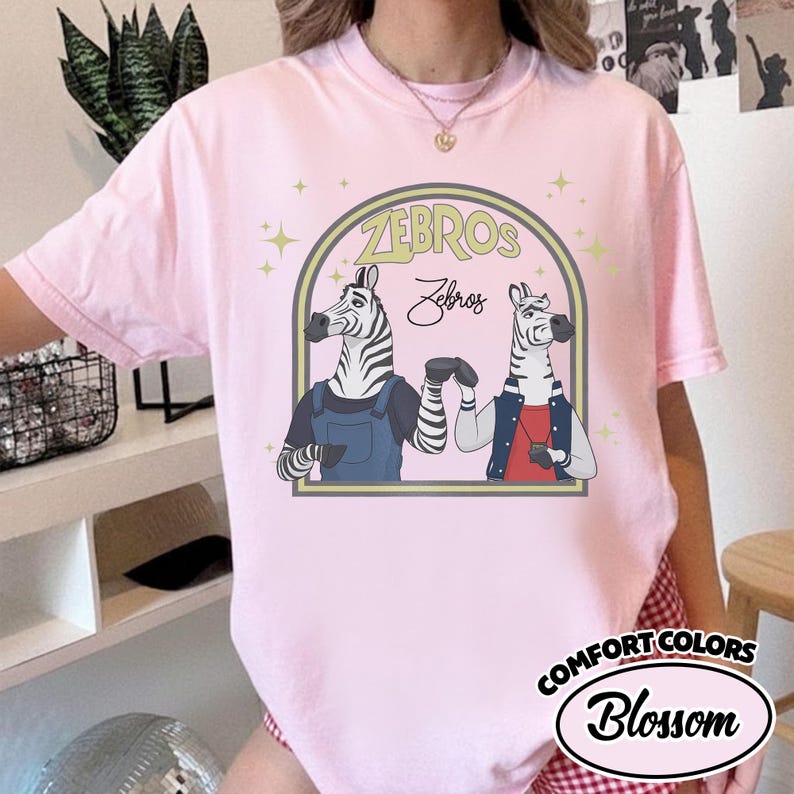 May include: A pale pink t-shirt with a cartoon illustration of two zebras inside an arch. The zebras are dressed in outfits, with the word "ZEBROS" above them. The shirt also has the text "Comfort Colors Blossom".
