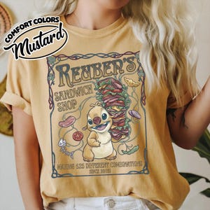 K&ouml;nnte beinhalten: Senffarbenes T-Shirt mit einer Cartoonfigur, die ein gro&szlig;es Sandwich h&auml;lt. Das Shirt zeigt "Reuber's Sandwich Shop" und "Making 625 Different Combinations Since 2008" in einem Vintage-Design.