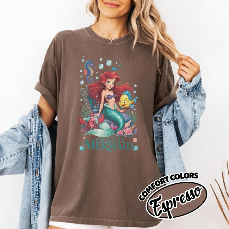 May include: Brown t-shirt featuring a colorful graphic of Ariel from The Little Mermaid, with her friends. The design includes the text "THE LITTLE MERMAID". The shirt also has the text "COMFORT COLORS Espresso".