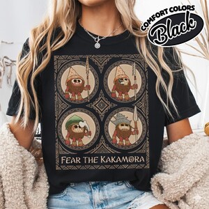 May include: Black t-shirt with a graphic of four cartoon figures with spears and hats, in circular frames and a decorative border. The text "FEAR THE KAKAMORA" is printed below. The words "COMFORT COLORS Black" are printed in a circular design at the top.