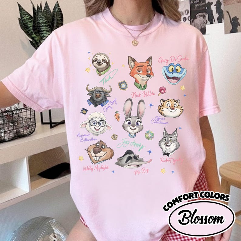 K&ouml;nnte beinhalten: Ein hellrosa T-Shirt mit Cartoon-Gesichtern von Zootopia. Das Shirt enth&auml;lt den Text "COMFORT COLORS" und "Blossom" in einem kreisf&ouml;rmigen Design. Das T-Shirt ist ein Rundhals-Stil.