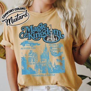 May include: Mustard-colored t-shirt featuring a blue graphic of "Magic Kingdom" with Cinderella Castle, Autopia Car, and Peter Pan's Flight. The shirt has rolled-up sleeves and a "Comfort Colors" logo.