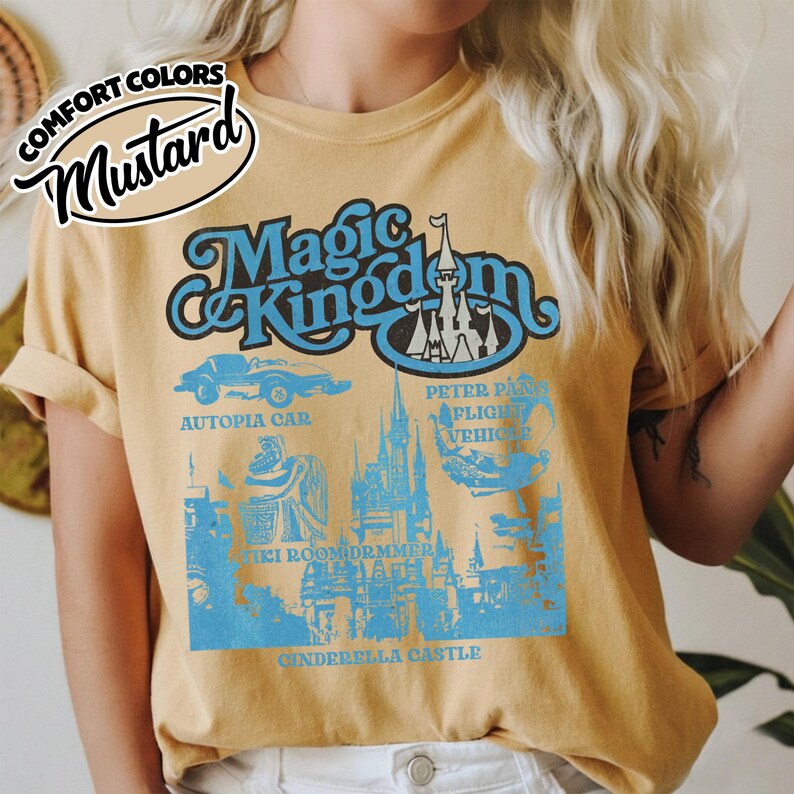 May include: Mustard-colored t-shirt featuring a blue graphic of "Magic Kingdom" with Cinderella Castle, Autopia Car, and Peter Pan's Flight. The shirt has rolled-up sleeves and a "Comfort Colors" logo.
