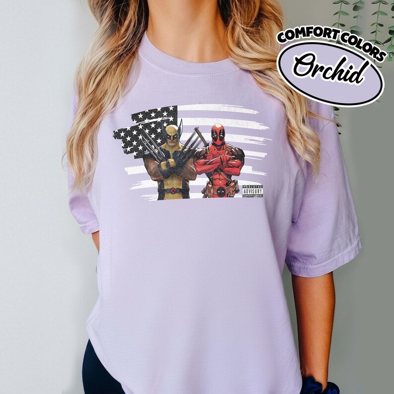 May include: A light purple t-shirt featuring a graphic print of Wolverine and Deadpool standing in front of a stylized American flag. The design includes a small black and white Parental Advisory label and the text Comfort Colors Orchid.