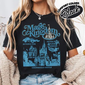 May include: Black t-shirt with a blue graphic design featuring the words "Magic Kingdom" and illustrations of a car, Cinderella Castle, and other attractions. The shirt also has the words "Comfort Colors" and "Black" in a circular design.