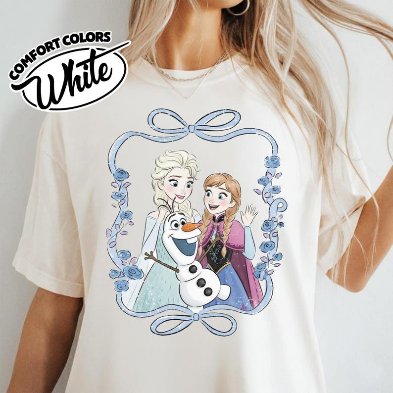 May include: White Comfort Colors t-shirt featuring a cartoon illustration of Elsa, Anna, and Olaf from the movie Frozen. The design is framed with a light blue bow and floral elements. The shirt has the word "White" in a cursive font.