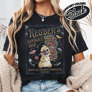 K&ouml;nnte beinhalten: Schwarzes T-Shirt mit einer Cartoonfigur, die ein gro&szlig;es Sandwich h&auml;lt. Das Shirt zeigt den Text "Reuben's Sandwich Shop" und "Making 625 Different Combinations Since 2003!" in einem dekorativen Rahmen. Die Worte "Comfort Colors" und "Black" sind in einem Kreis.