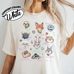 K&ouml;nnte beinhalten: Wei&szlig;es Comfort Colors T-Shirt mit Cartoon-Illustrationen von Charakteren aus einem beliebten Zeichentrickfilm. Das Shirt zeigt Charaktere wie Judy Hopps, Nick Wilde und Flash. Der Text "COMFORT COLORS White" ist oben links aufgedruckt.