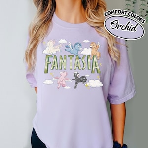 May include: A lilac t-shirt with the word "FANTASIA" in green. The design includes cartoon pegasus characters in white, blue, pink, and black, with clouds and stars. The text "COMFORT COLORS" and "Orchid" are in a white oval.