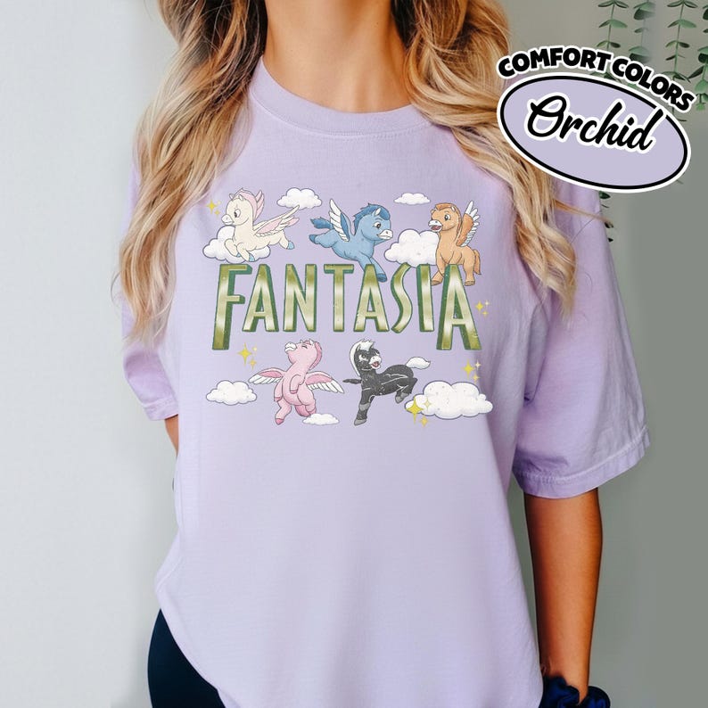 May include: A lilac t-shirt with the word "FANTASIA" in green. The design includes cartoon pegasus characters in white, blue, pink, and black, with clouds and stars. The text "COMFORT COLORS" and "Orchid" are in a white oval.