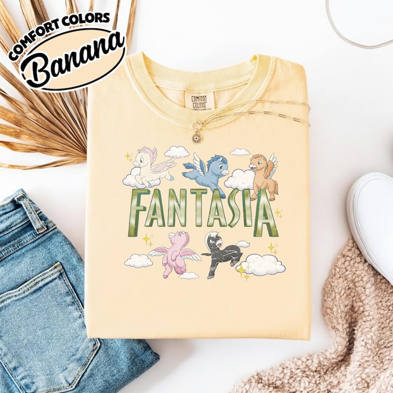 May include: A pale yellow t-shirt with the word "FANTASIA" in green. The shirt features cartoon pegasus characters in various colours, including white, blue, and pink. The shirt has the words "COMFORT COLORS" and "Banana" in a circular design.