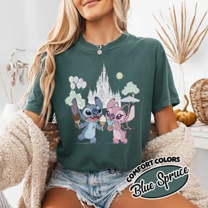 Puede incluir: Camiseta Blue Spruce Comfort Colors con una ilustraci&oacute;n de dibujos animados de Stitch y Angel sosteniendo conos de helado frente a un castillo. El dise&ntilde;o incluye globos, &aacute;rboles y un sol. La camiseta tiene las palabras "Comfort Colors" y "Blue Spruce".