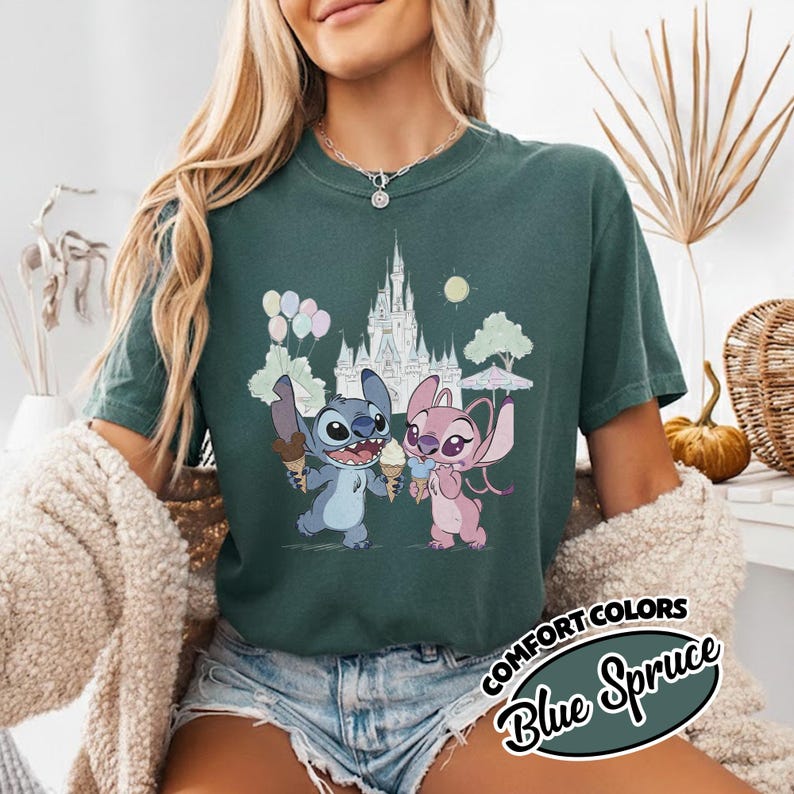 Puede incluir: Camiseta Blue Spruce Comfort Colors con una ilustraci&oacute;n de dibujos animados de Stitch y Angel sosteniendo conos de helado frente a un castillo. El dise&ntilde;o incluye globos, &aacute;rboles y un sol. La camiseta tiene las palabras "Comfort Colors" y "Blue Spruce".