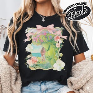 K&ouml;nnte beinhalten: Schwarzes T-Shirt mit einer floralen und Gesichts-Grafik. Die Grafik zeigt ein Frauengesicht mit Blumenkrone, einer rosa Schleife und einem Paar am Strand. Die Worte "COMFORT COLORS Black" sind oben rechts aufgedruckt.
