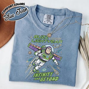 May include: A blue jean colored t-shirt featuring a graphic of Buzz Lightyear flying through space with the text Buzz Lightyear to infinity and beyond. The shirt has a crew neckline and a Comfort Colors brand label on the inside of the collar.
