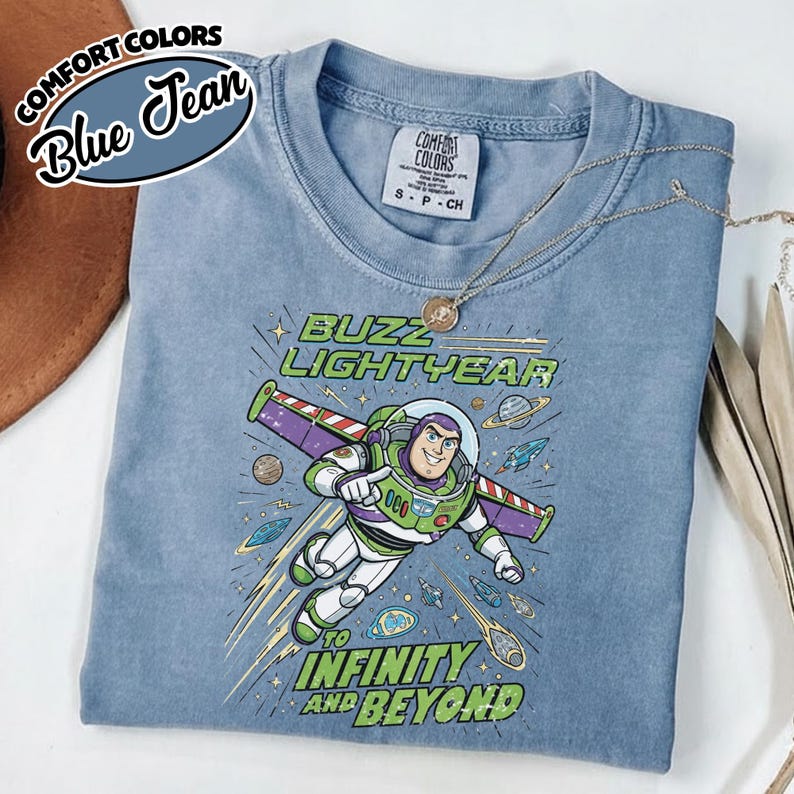 May include: A blue jean colored t-shirt featuring a graphic of Buzz Lightyear flying through space with the text Buzz Lightyear to infinity and beyond. The shirt has a crew neckline and a Comfort Colors brand label on the inside of the collar.