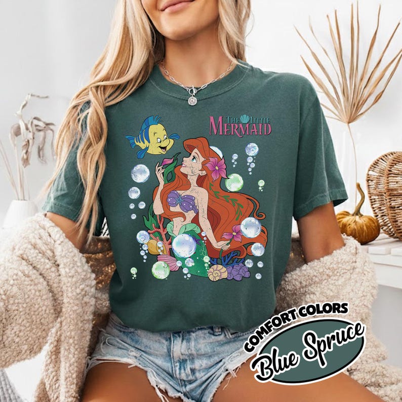 K&ouml;nnte beinhalten: Ein dunkelgr&uuml;nes T-Shirt mit einem Grafikdruck von Arielle, die Meerjungfrau, mit rotem Haar, violettem Muschel-Oberteil und gr&uuml;nem Schwanz. Der Fisch Fabius ist daneben, umgeben von Blasen, Seetang und dem Schriftzug The Little Mermaid.