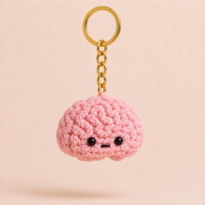 May include: A pink, crocheted brain-shaped keychain with black eyes and a small mouth. The keychain has a gold-colored ring and chain. The background is a soft pink.