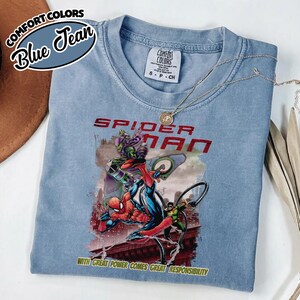 Op de afbeelding: Lichtblauw T-shirt met een Spider-Man-afbeelding. De afbeelding bevat de rode woorden "SPIDER MAN" en de zin "WITH GREAT POWER COMES GREAT RESPONSIBILITY". Het shirt heeft de woorden "COMFORT COLORS" en "Blue Jean" in een cirkelvormig ontwerp.