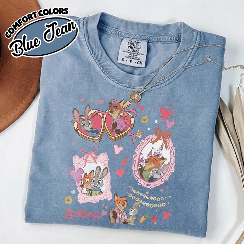 May include: A blue jean colored t-shirt featuring a graphic print of Zootopia characters Judy Hopps and Nick Wilde. The design includes heart-shaped and ornate frames, pink bows, small hearts, and Mickey Mouse ear silhouettes. The shirt has a Comfort Colors label.