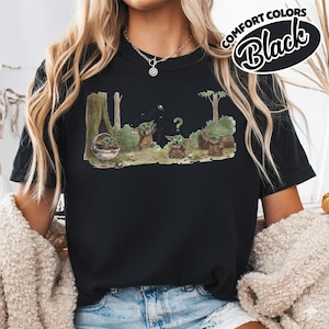 May include: Black t-shirt featuring a cartoon illustration of a green alien character in a forest setting. The graphic includes the text "COMFORT COLORS Black". The alien is depicted in various poses, with a question mark above one.
