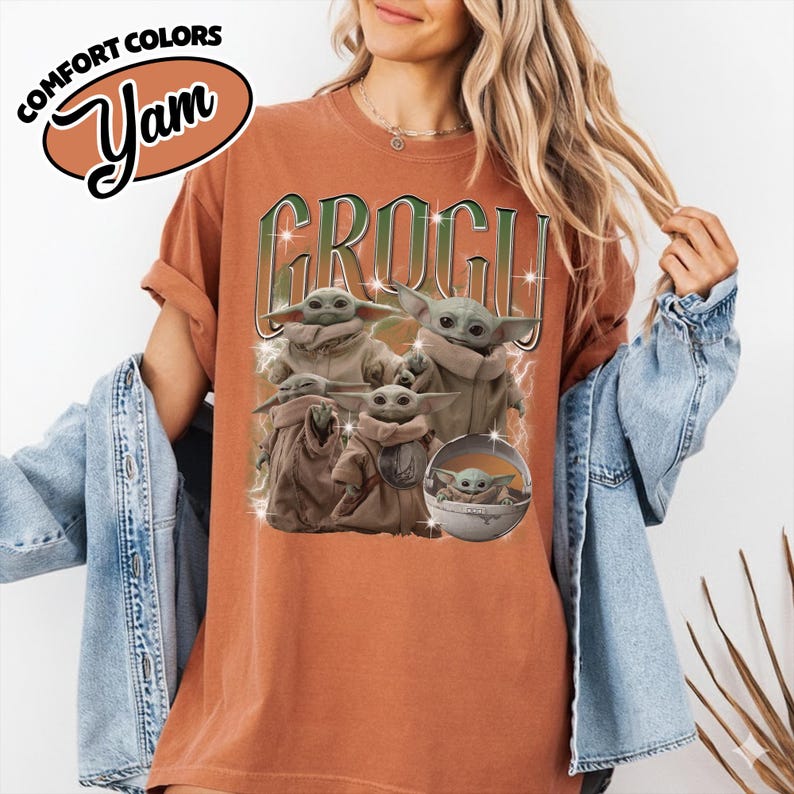 K&ouml;nnte beinhalten: Ein rostfarbenes Kurzarm-T-Shirt mit einem Grafikdruck von mehreren Grogu-Figuren. Der Text "GROGU" ist in gro&szlig;en gr&uuml;nen Buchstaben &uuml;ber den Figuren abgebildet. Ein Logo in der oberen linken Ecke zeigt "COMFORT COLORS Yam".