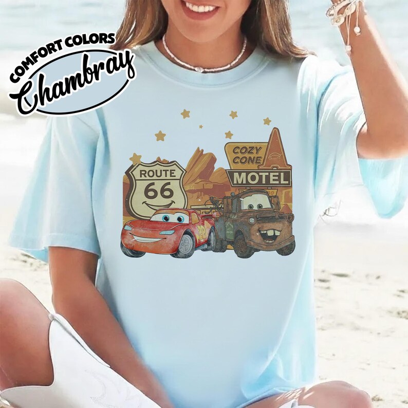 May include: Light blue t-shirt featuring a graphic of characters from the movie Cars, including Lightning McQueen and Mater, with a Route 66 sign and Cozy Cone Motel. The shirt has the words "Comfort Colors Chambray" in the upper left corner.
