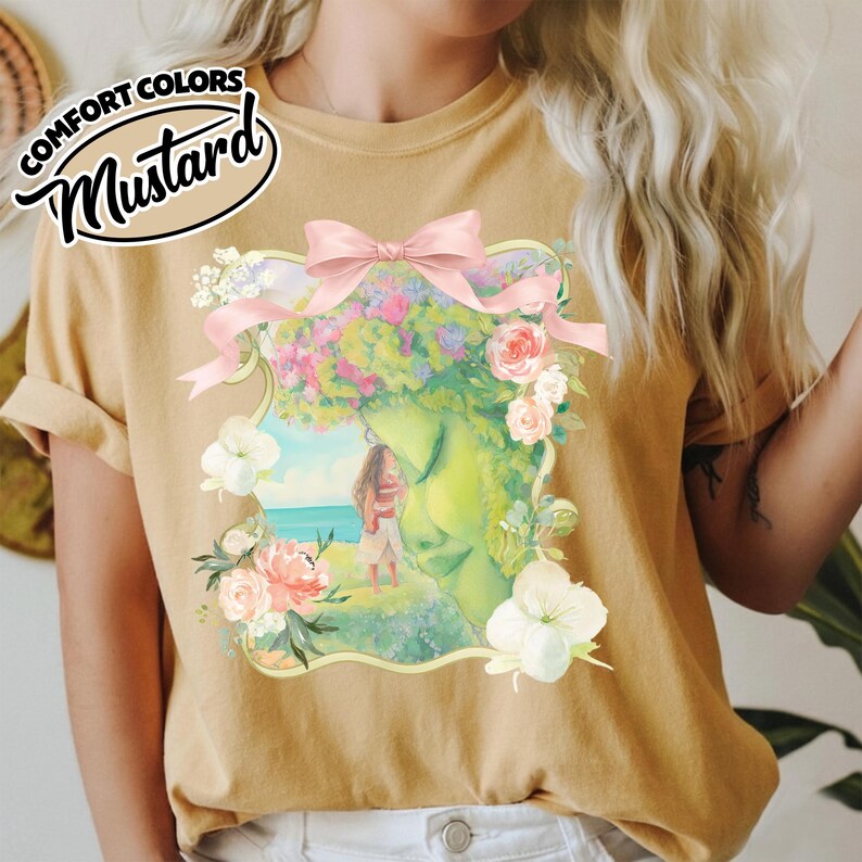 K&ouml;nnte beinhalten: Senffarbenes T-Shirt mit einer gerahmten Illustration, einer rosa Schleife und floralen Akzenten. Die Illustration zeigt eine Frau am Meer, mit gr&uuml;nem Gesicht und Blumenhaar. Der Text "COMFORT COLORS" und "Mustard" befindet sich oben links.