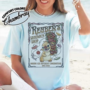 K&ouml;nnte beinhalten: Hellblaues T-Shirt mit einer Cartoon-Grafik, die eine Figur und ein Sandwich zeigt. Die Grafik zeigt "Reuber's Sandwich Shop" und "Making 625 Different Combinations Since 2003!". Das Shirt hat auch die Aufschrift "Comfort Colors Chambray".