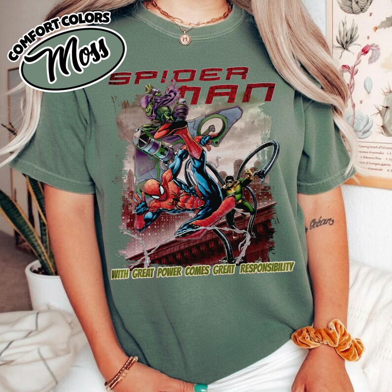 Op de afbeelding: Mosgroen T-shirt met een Spider-Man-afbeelding. Het ontwerp toont Spider-Man in actie met de Green Goblin en de tekst "SPIDER-MAN" en "WITH GREAT POWER COMES GREAT RESPONSIBILITY".