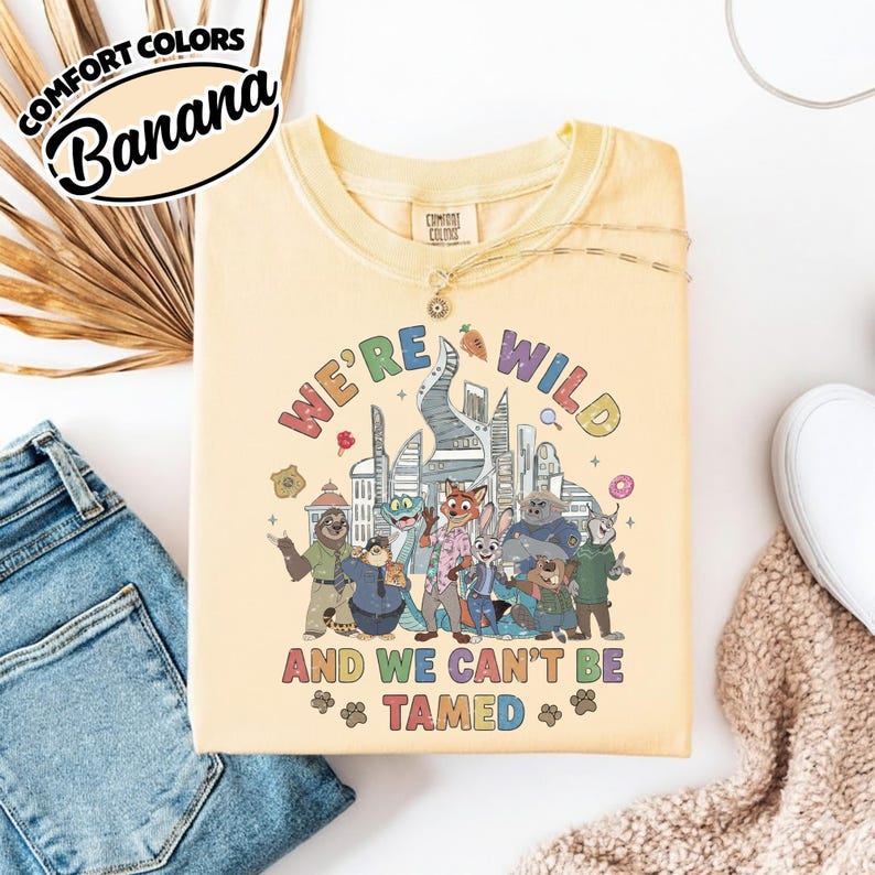 May include: A light yellow t-shirt with the text "WE'RE WILD AND WE CAN'T BE TAMED" and a colorful graphic of cartoon characters and a cityscape. The shirt has a crew neck and short sleeves.