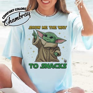 May include: Light blue t-shirt with the text "SHOW ME THE WAY TO SNACKS" and a graphic of a green alien character. The shirt has the words "COMFORT COLORS" and "Chambray" printed on the top left.