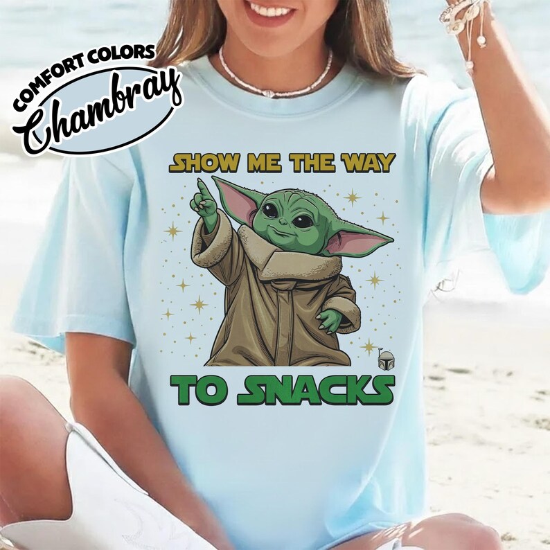 May include: Light blue t-shirt with the text "SHOW ME THE WAY TO SNACKS" and a graphic of a green alien character. The shirt has the words "COMFORT COLORS" and "Chambray" printed on the top left.