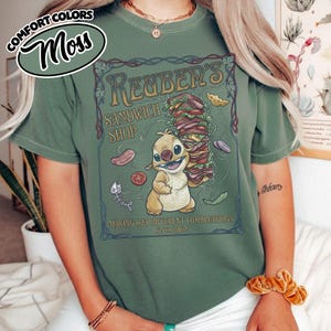 K&ouml;nnte beinhalten: Moosgr&uuml;nes T-Shirt mit einer Cartoonfigur, die ein gro&szlig;es Sandwich h&auml;lt. Das Shirt tr&auml;gt den Text "Reuben's Sandwich Shop" und "Making 625 Different Combinations Since 2002". Auf dem Shirt stehen die Worte "Comfort Colors" in einem kreisf&ouml;rmigen Design.