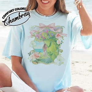 K&ouml;nnte beinhalten: Hellblaues T-Shirt mit einer floralen und fig&uuml;rlichen Grafik. Die Grafik zeigt das Gesicht einer Frau mit Blumenkrone, einer rosa Schleife und einer Strandszene. Der Text "COMFORT COLORS Chambray" ist oben links aufgedruckt.