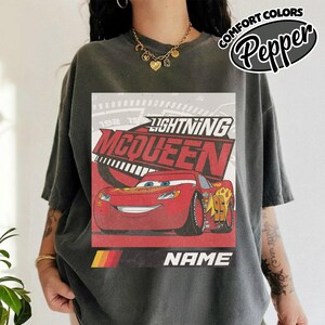 May include: A charcoal gray short-sleeve t-shirt featuring a graphic of the red race car Lightning McQueen. The design includes the text Lightning McQueen, the number 95, and a placeholder for a name. The shirt has a Comfort Colors label.