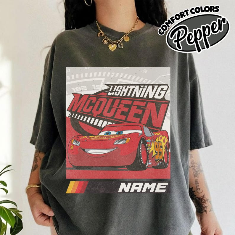 May include: A charcoal gray short-sleeve t-shirt featuring a graphic of the red race car Lightning McQueen. The design includes the text Lightning McQueen, the number 95, and a placeholder for a name. The shirt has a Comfort Colors label.
