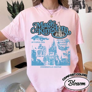 May include: A light pink t-shirt with a blue graphic featuring the words "Magic Kingdom" and illustrations of an Autopia car, Cinderella Castle, and other theme park attractions. The shirt also has the words "Comfort Colors" and "Blossom".
