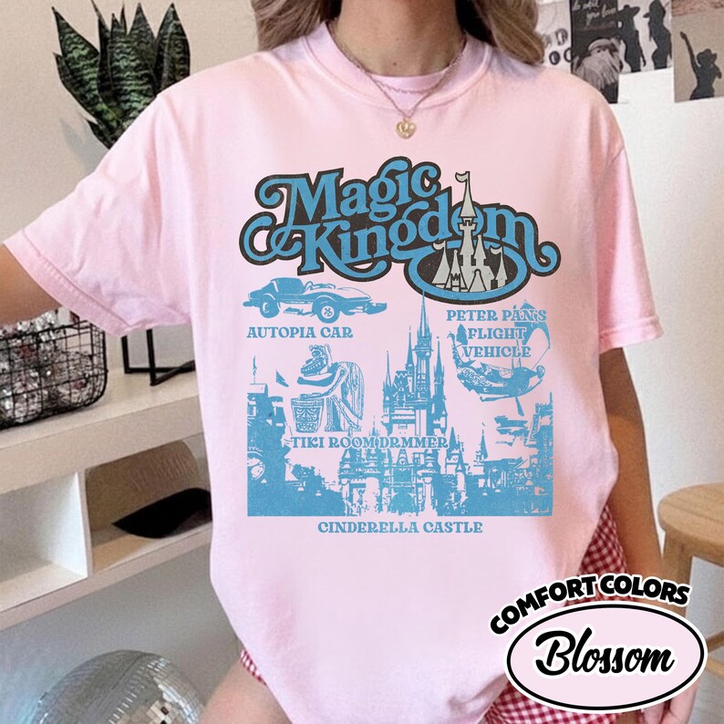 May include: A light pink t-shirt with a blue graphic featuring the words "Magic Kingdom" and illustrations of an Autopia car, Cinderella Castle, and other theme park attractions. The shirt also has the words "Comfort Colors" and "Blossom".