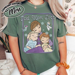 May include: A moss green t-shirt featuring the phrase "Best Mom Ever" with a graphic of a mother and two children, along with cartoon characters and snowflakes. The shirt has a relaxed fit and is made of a soft material.