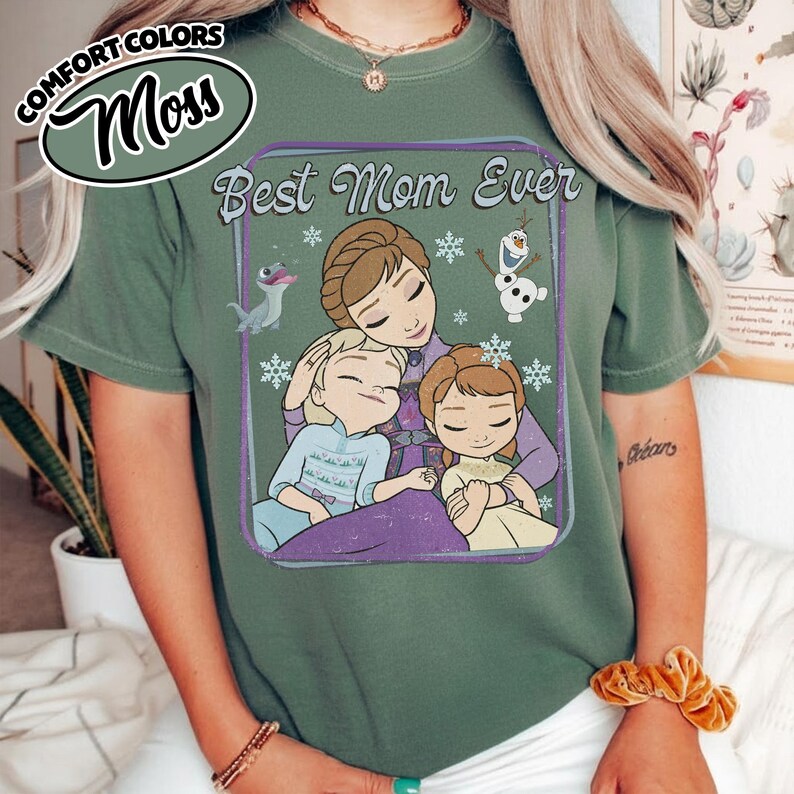 May include: A moss green t-shirt featuring the phrase "Best Mom Ever" with a graphic of a mother and two children, along with cartoon characters and snowflakes. The shirt has a relaxed fit and is made of a soft material.