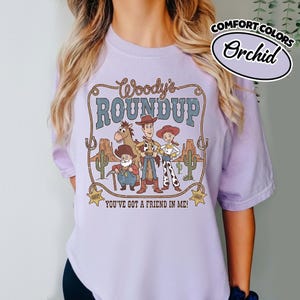 May include: Orchid-colored t-shirt featuring a graphic from "Woody's Roundup." The design includes cartoon characters, cacti, and the text "Woody's Roundup" and "You've Got a Friend in Me!" The shirt is labeled "Comfort Colors."