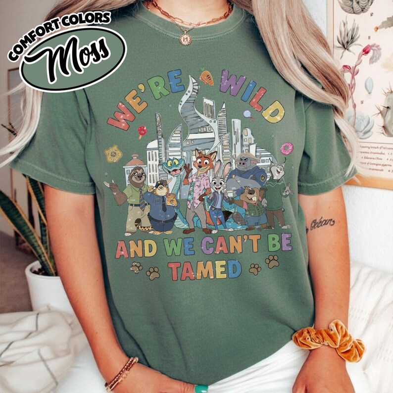 May include: Moss green t-shirt with the text "WE'RE WILD AND WE CAN'T BE TAMED" and a colorful graphic of animated animal characters. The shirt has short sleeves and a crew neck.