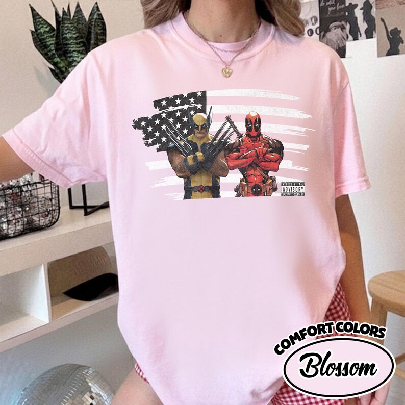 May include: A light pink t-shirt featuring a graphic print of Wolverine and Deadpool standing in front of a distressed American flag. The graphic includes a small black and white Parental Advisory Explicit Content label in the bottom right corner.
