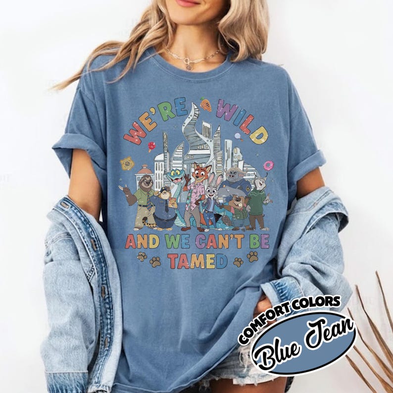 May include: Blue Comfort Colors t-shirt with the text "WE'RE WILD" and "AND WE CAN'T BE TAMED" in colorful letters. The shirt features a cartoon illustration of various animal characters in front of a city skyline.